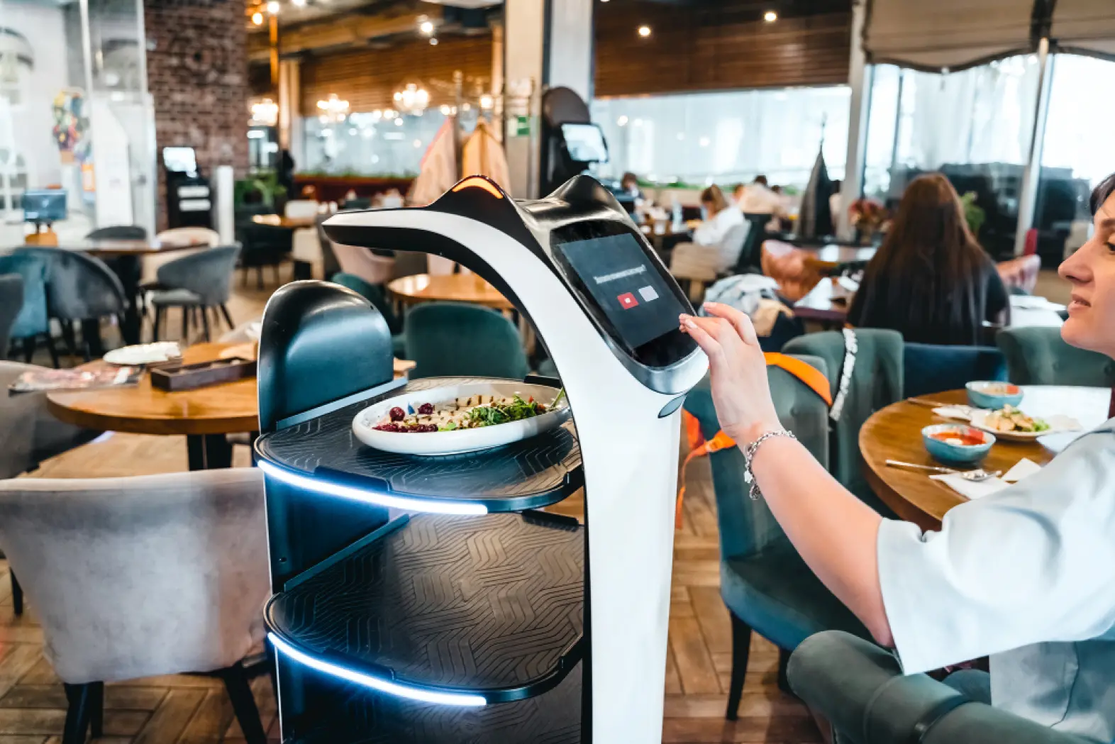 A person uses a touchscreen on a robot server delivering food in a modern restaurant, with patrons seated at tables in the background.